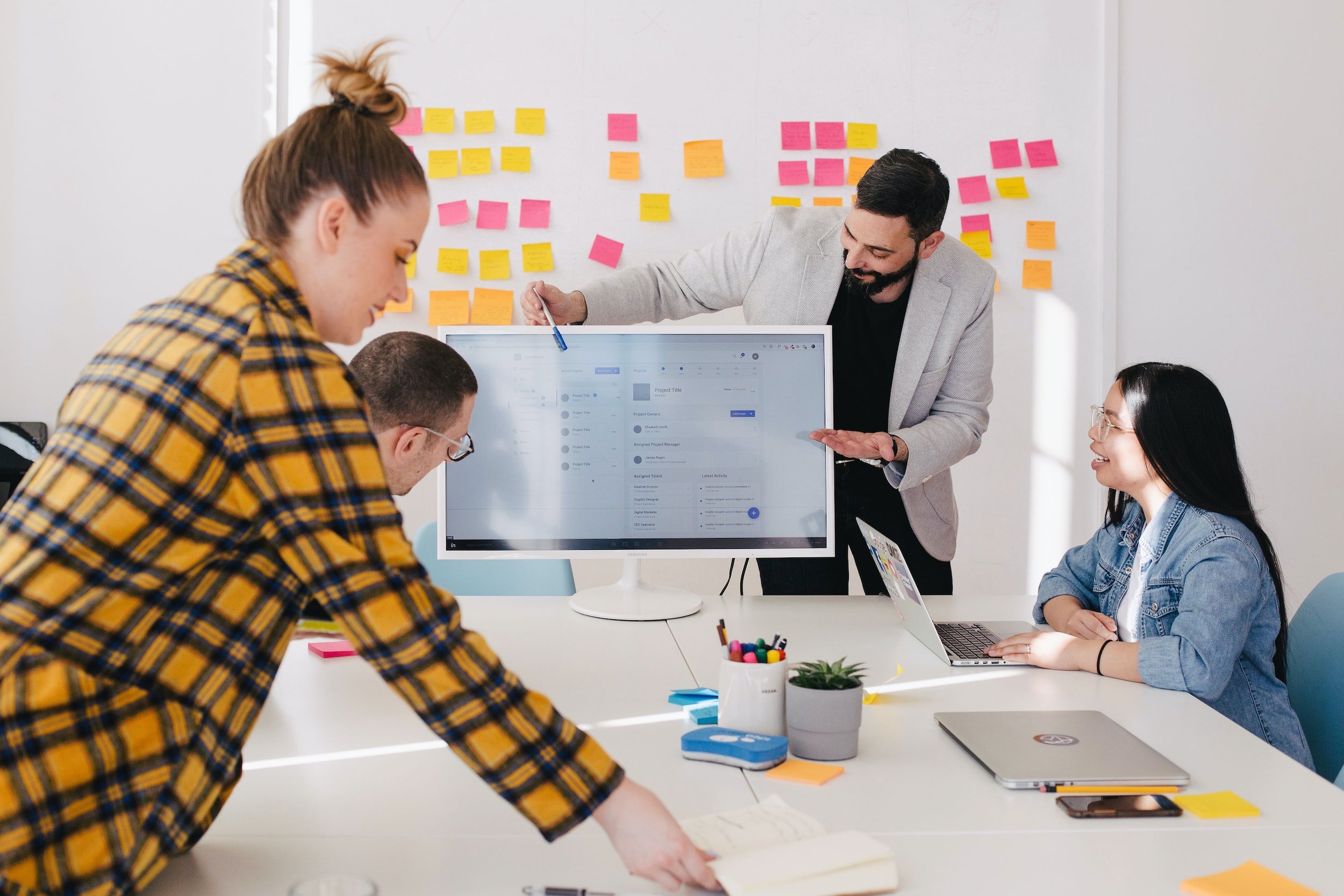 Business meeting with post-its on the wall and a man pointing to computer screen with pen to the rest of the team